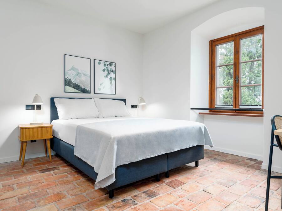 Une chambre à coucher avec un lit, deux tables de chevet avec des lampes, une fenêtre donnant sur des arbres et un plancher en briques.