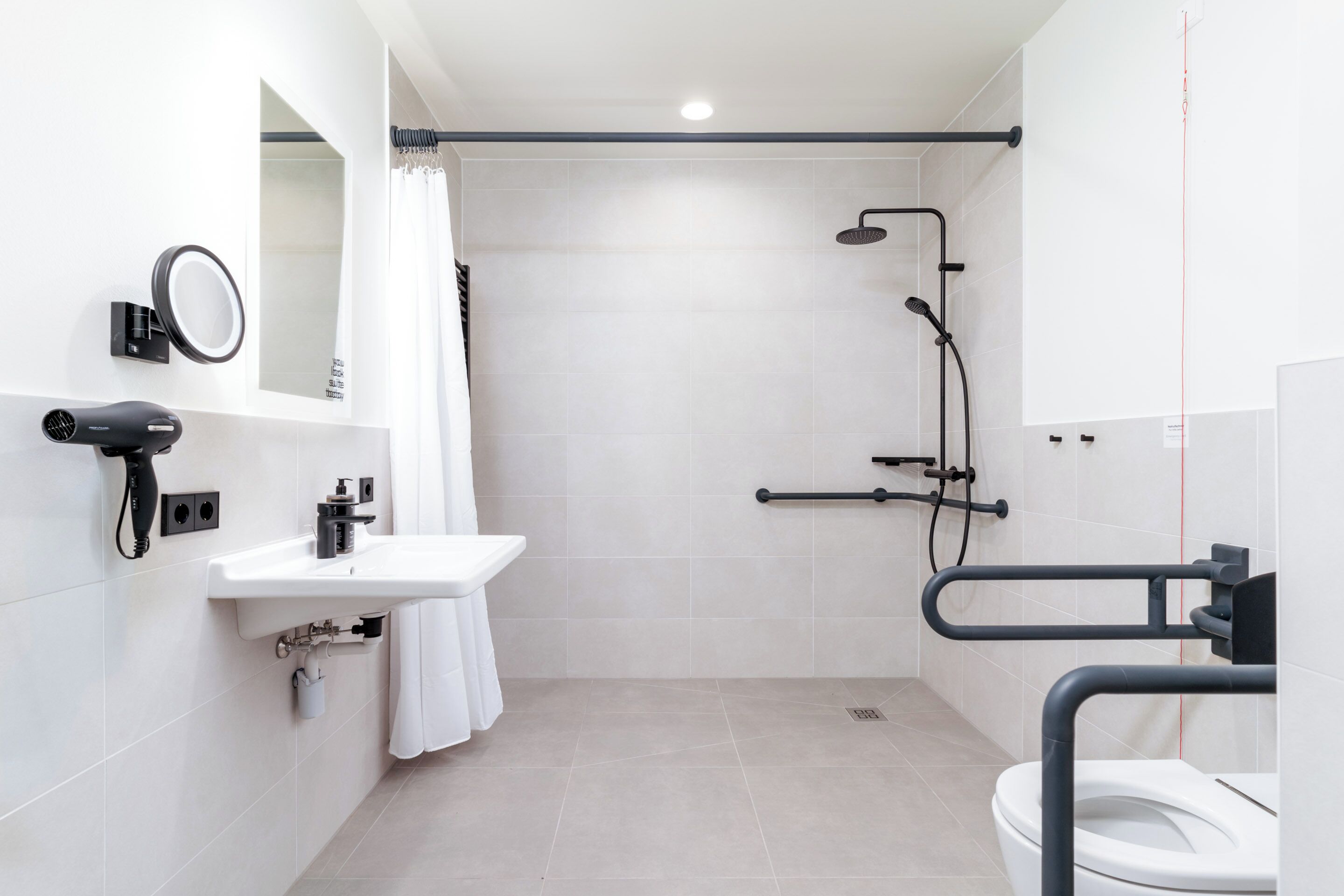 Ein modernes Badezimmer mit Waschbecken, Spiegel und Toilette. Es gibt eine Duschkabine mit Duschbrause und Haltegriff.