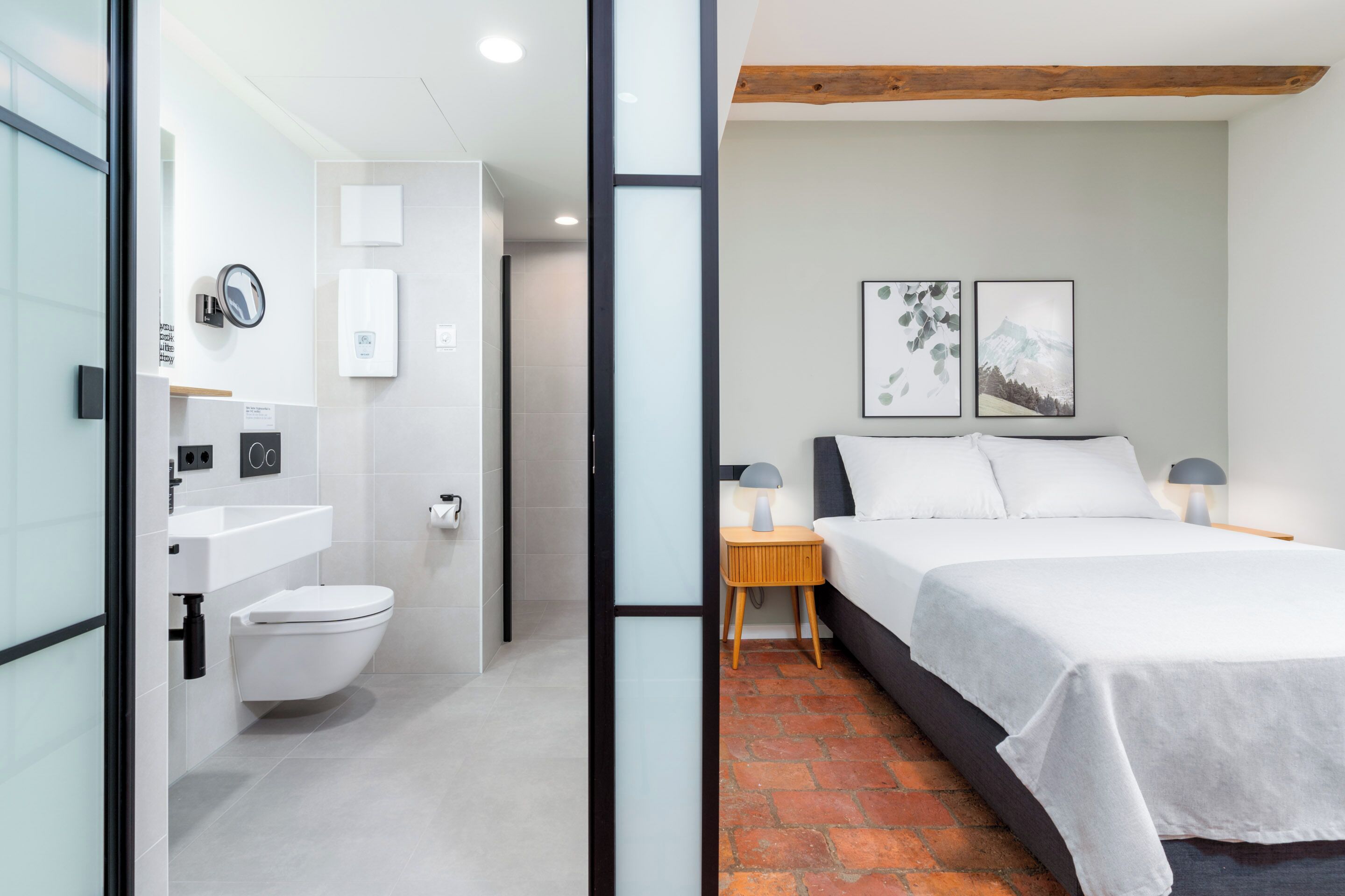 A modern hotel room with a white toilet, a sink, a bedside table with a lamp, a bed with white bedding, and two framed pictures on the wall.