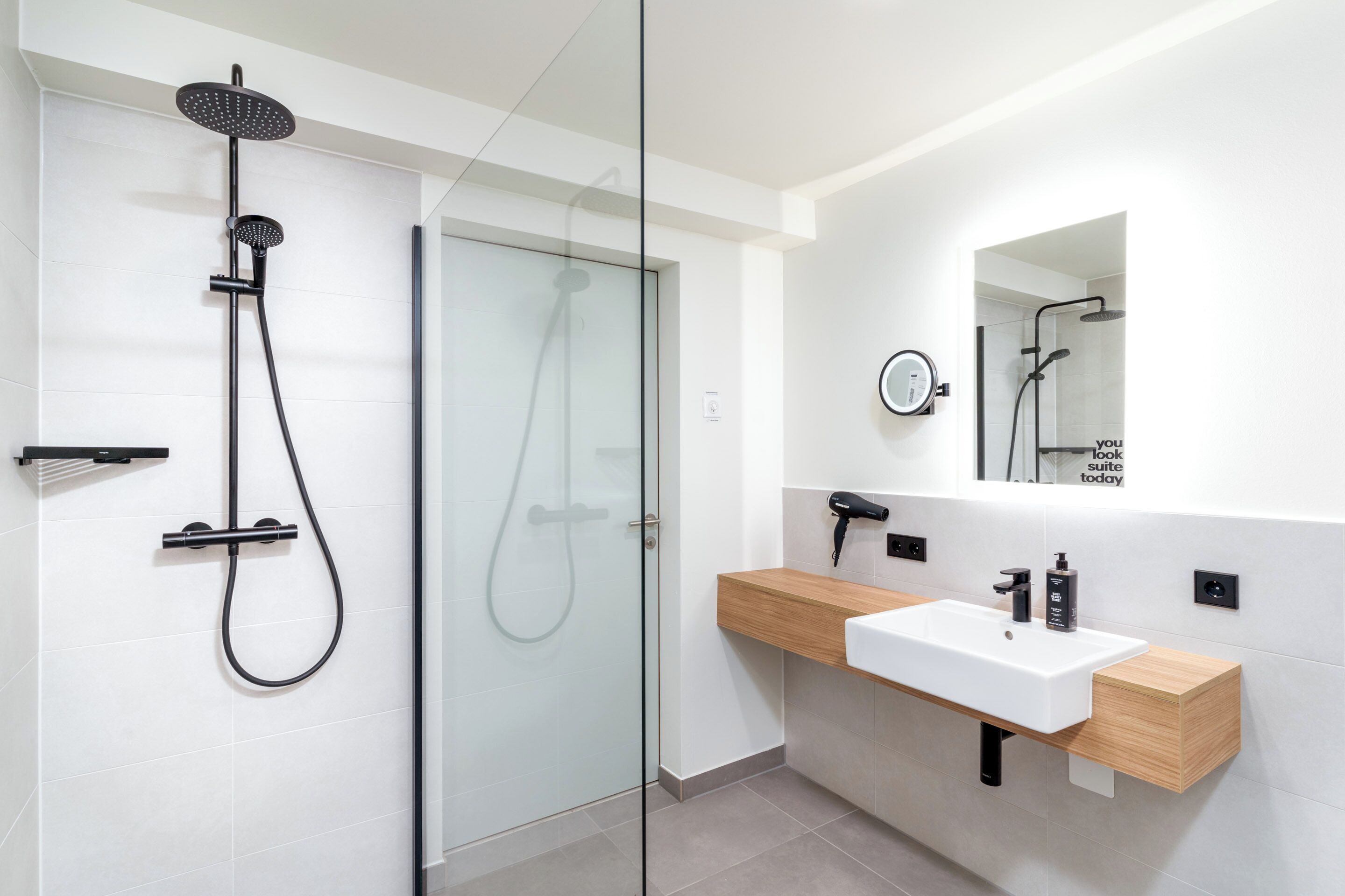 Une salle de bain moderne dotée d’une douche à parois de verre, d’un meuble-lavabo en bois et d’un miroir orné d’une citation inspirante.