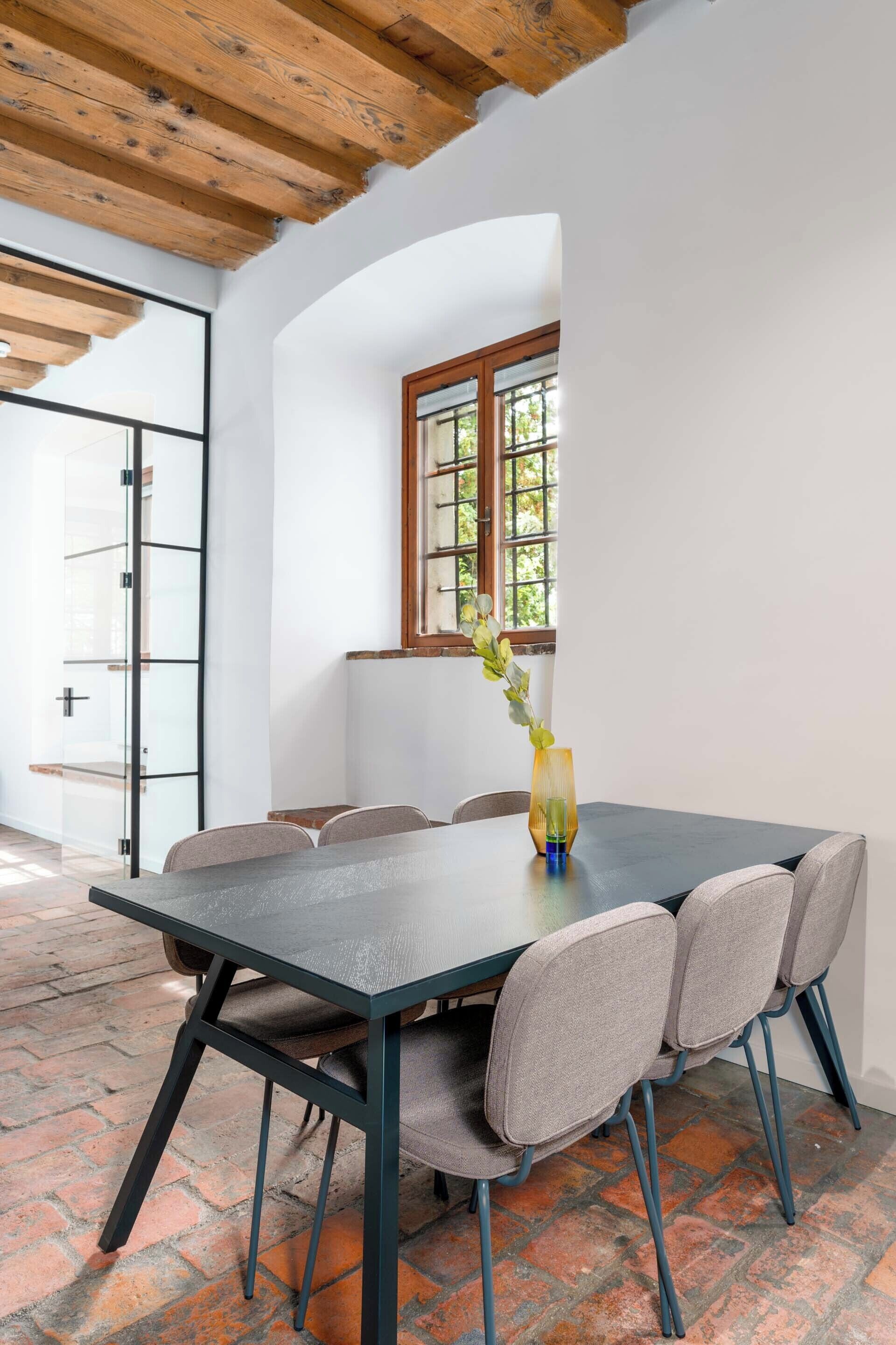 A dining area with a table and chairs, a vase with a plant, and a window with a view of greenery.