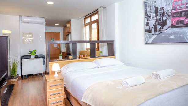 A hotel room with a bed, a desk, a mirror, a window, and a painting of a bus. - Apartamentos Turísticos Distrito Romano (Cartagena)