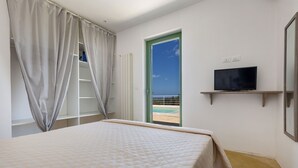 Une chambre à coucher avec un lit, un téléviseur posé sur une étagère, une fenêtre avec des rideaux et une vue sur la plage.