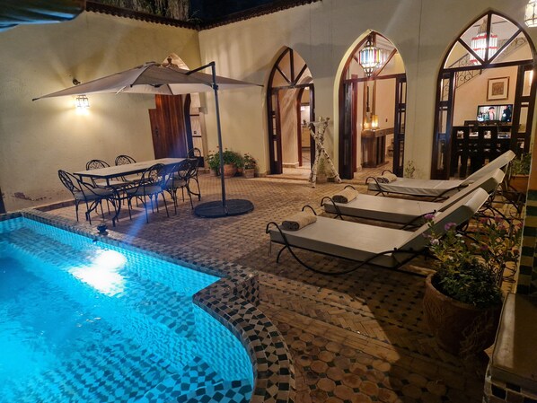Un espace extérieur avec piscine, doté d’un sol carrelé, de chaises longues, d’une table avec des chaises et de parasols.