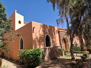 Un bâtiment doté de fenêtres en arc de cercle et d’un toit plat, entouré de verdure et surplombé d’un ciel dégagé.
