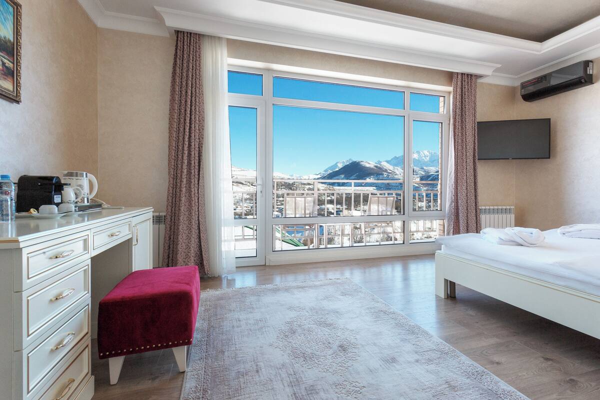 A hotel room with a large window, a bed, a desk with a coffee maker, and a TV.