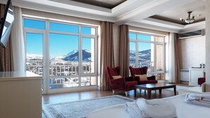 Habitación de hotel con una cama grande, dos sillones rojos, una mesa de centro de madera y vistas a montañas nevadas.