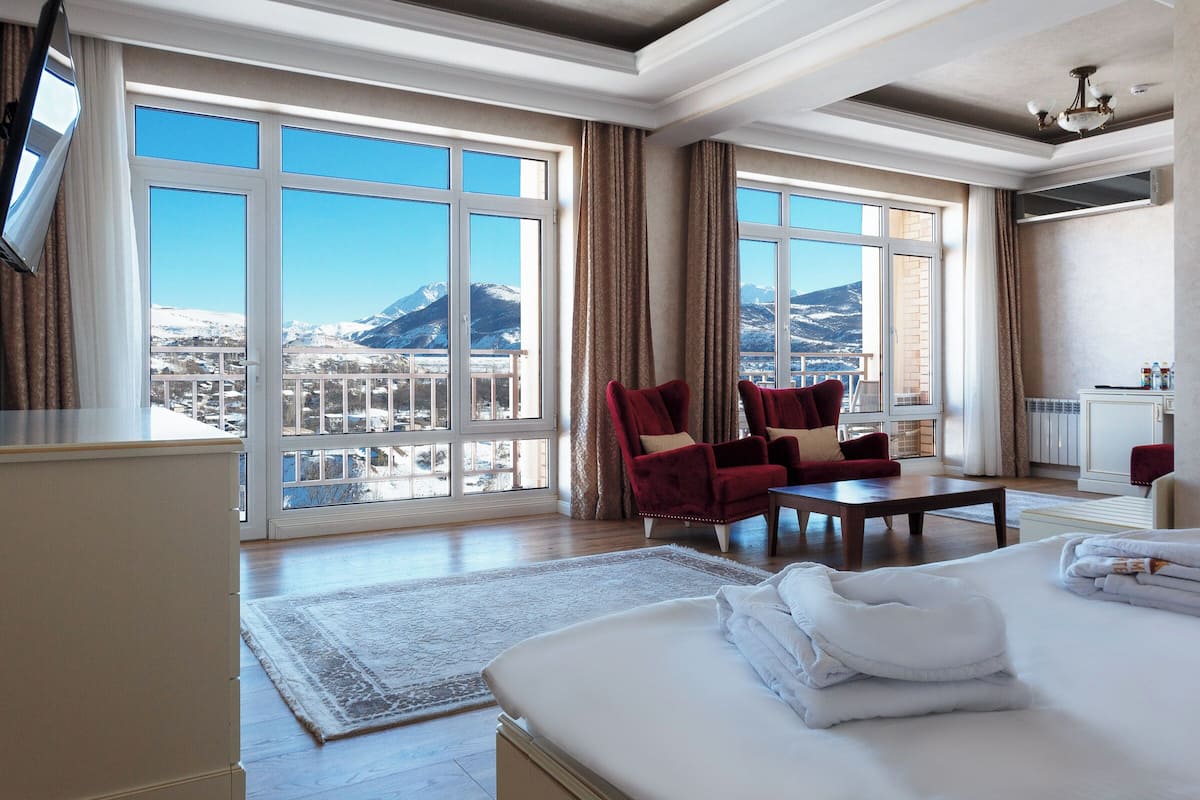 A hotel room with a large bed, two red armchairs, a wooden coffee table, and a view of snow-capped mountains.