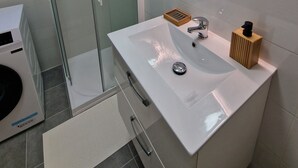 A modern bathroom with a glass-enclosed shower, a white sink with a wooden soap dispenser, and a large oval mirror.