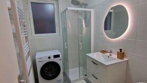 A compact bathroom with a washing machine, a glass-enclosed shower, a toilet, and a sink with a mirror.