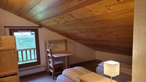 A small attic bedroom with a sloped ceiling, a window with a view of greenery, a wooden chair, a bed with a white comforter, and a bedside lamp. - Spacious T4 with garage and parking in Grand-Bornand - 6 pers (Le Grand-Bornand)