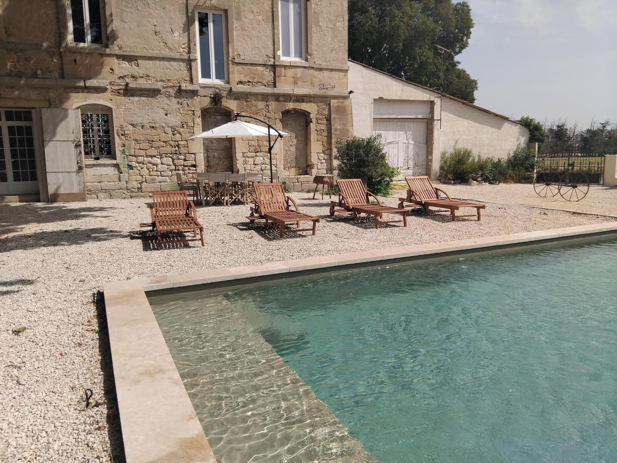 Un área de piscina con tumbonas de madera y una mesa con sillas, rodeada por un patio de grava y un edificio de piedra con ventanas.