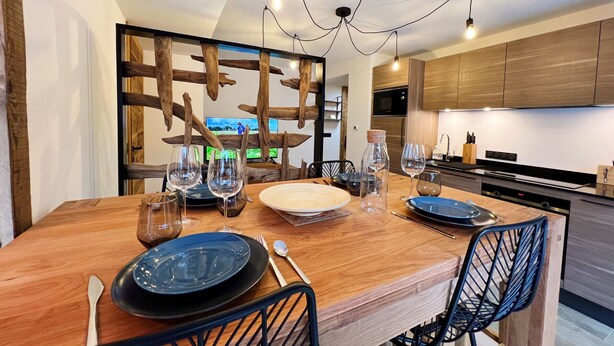 A modern dining area with a wooden table set for four, surrounded by black chairs. A television is mounted on a wooden partition, and the kitchen area is visible in the background. - Charming apartment in Les Gets, 2 bedrooms, Wi-Fi, parking included (Les Gets)