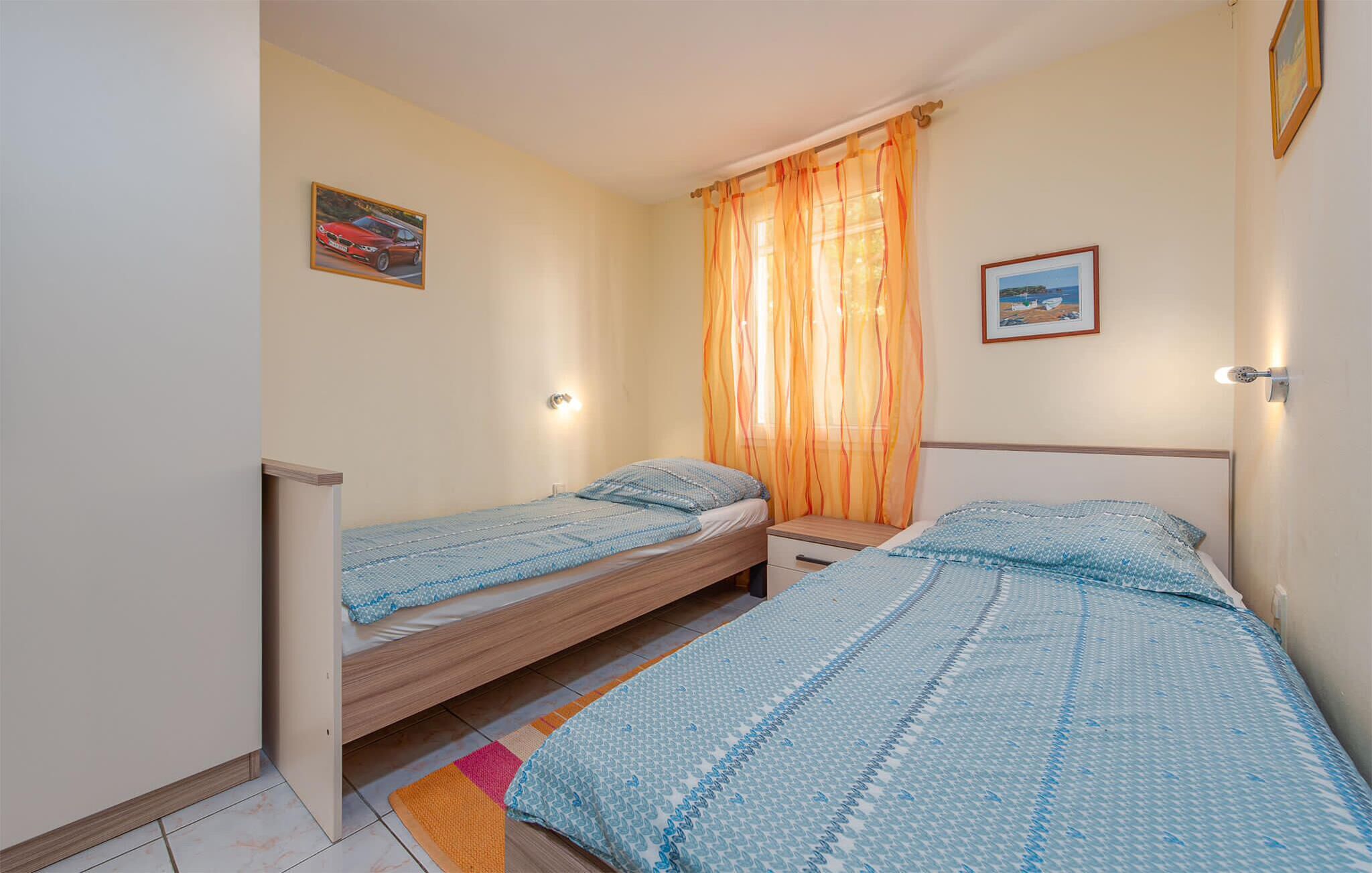 Un dormitorio pequeño y bien iluminado con dos camas individuales, una mesita de noche, una ventana con cortinas translúcidas y una lámpara fijada a la pared.