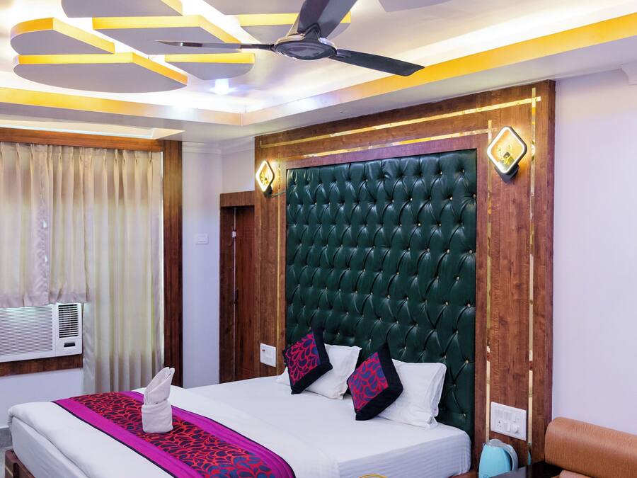 Habitación de hotel con una cama grande, cabecera de madera, mesita de noche con lámpara, sofá y ventilador de techo.