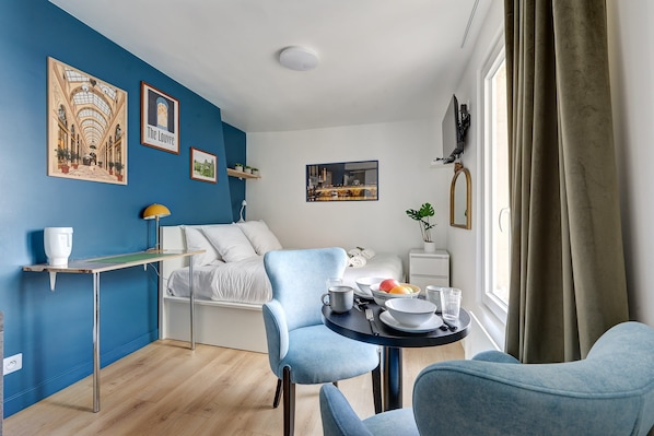 Quarto de hotel moderno com parede azul, cama com roupa de cama branca, área de refeições com uma cadeira azul, uma pequena mesa com uma tigela de frutas e uma janela com cortinas.