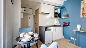 A compact kitchen with white cabinets, a microwave, and a small refrigerator. There is a dining table with chairs and a fruit bowl. - Studio de charme au cœur de Montorgueil (Paris)