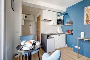 Une cuisine compacte avec des meubles blancs, un four à micro-ondes et un petit réfrigérateur. On y trouve une table à manger avec des chaises et un panier de fruits.