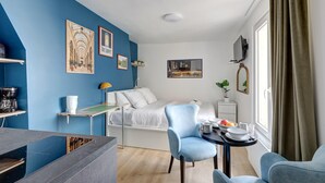 A modern room with a blue accent wall, a bed with white bedding, a dining area with a table and chairs, and a TV mounted on the wall. - Studio de charme au cœur de Montorgueil (Paris)