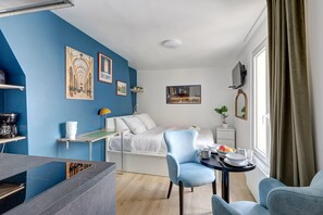 Une chambre moderne avec un mur d’accent bleu, un lit avec une literie blanche, un coin repas avec une table et des chaises, et un téléviseur fixé au mur.