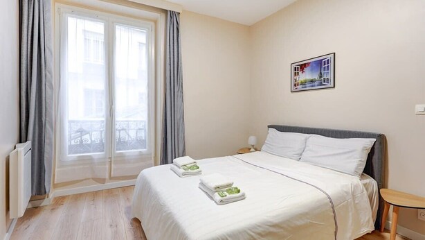 Habitación de hotel con una cama grande, una mesita de noche pequeña, una ventana con cortinas y un cuadro en la pared.