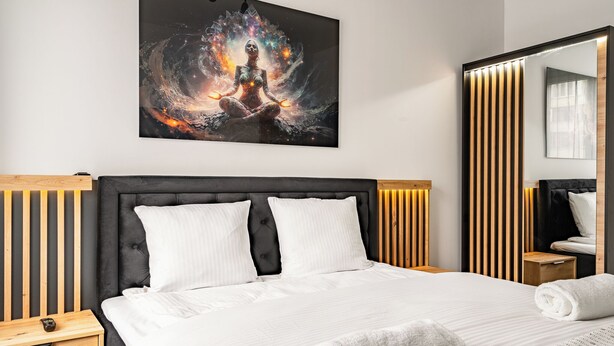 A modern hotel room with a large bed, a nightstand, a wooden headboard, and a wall-mounted artwork depicting a meditative figure. - RentPlanet - Apartament Grabiszyńska (Wroclaw)