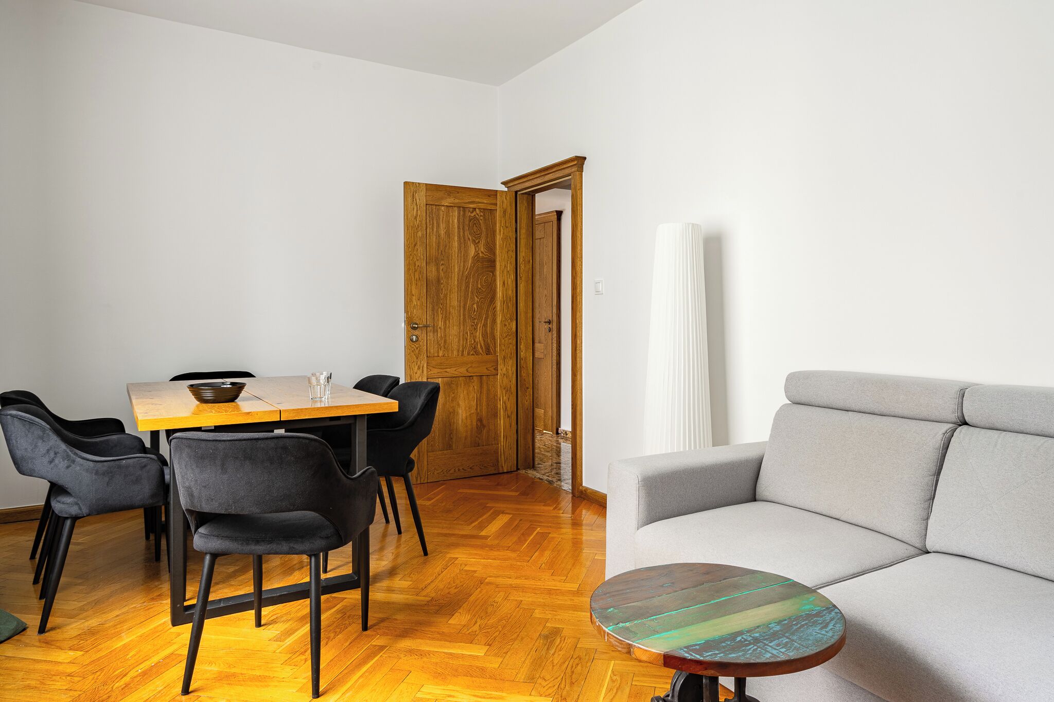 A dining area with a wooden table and black chairs, a grey sofa, and a herringbone wooden floor.