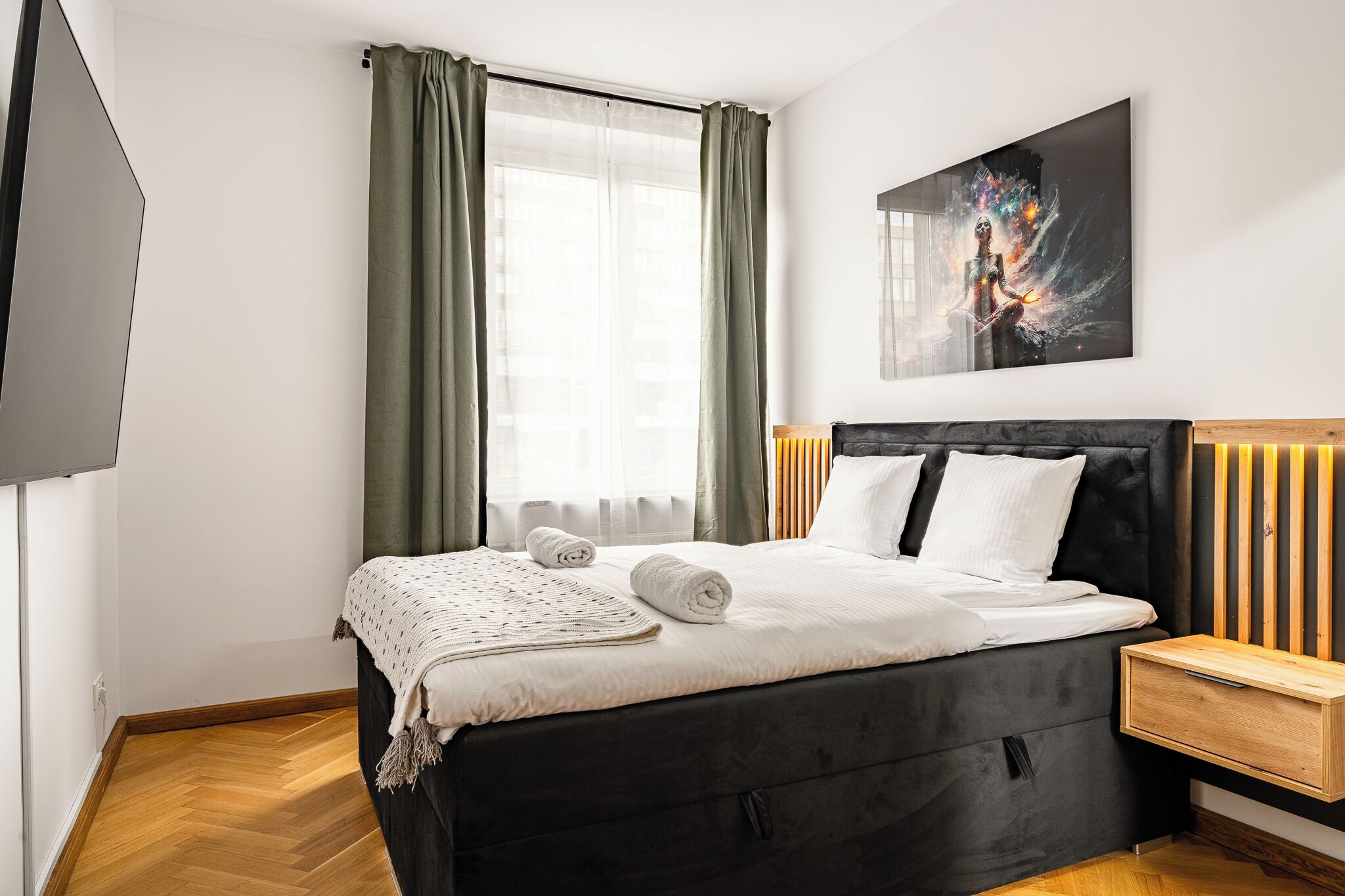 A modern bedroom with a black bed, white bedding, a wooden nightstand, and a wall-mounted TV.