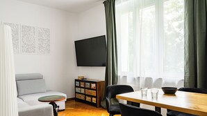 A modern living room with a grey sofa, a wooden dining table, black chairs, a flat-screen TV, and a cabinet with decorative items. - RentPlanet - Apartament Grabiszyńska (Wroclaw)