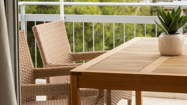 A balcony with wicker chairs and a wooden table, a potted plant, and a view of buildings and trees. - Beachfront 2-bedroom Stay in Voula (Vari-Voula-Vouliagmeni)