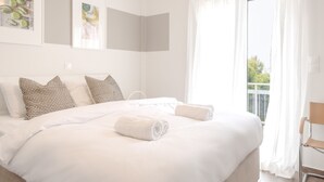 A neatly made bed with white linens and two rolled towels, a large window with sheer curtains, and a wall-mounted air conditioning unit. - Beachfront 2-bedroom Stay in Voula (Vari-Voula-Vouliagmeni)