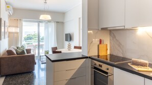 A modern kitchen with a black countertop, white cabinets, and a built-in oven. There is a dining area with a table and chairs, and a sofa in the background. - Beachfront 2-bedroom Stay in Voula (Vari-Voula-Vouliagmeni)
