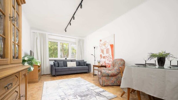 A spacious living room with a grey sofa, a floral armchair, a dining table with a white tablecloth, and a large window with curtains.