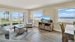 A spacious living room with a large TV, comfortable seating, and a view of the beach.
