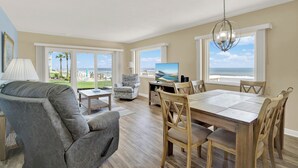 A spacious living room with a large window overlooking the ocean, a comfortable gray sofa, a wooden dining table with chairs, and a television on a stand.