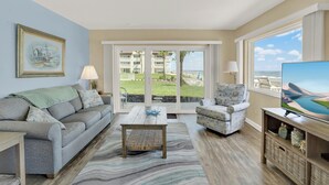 A living room with a sofa, armchair, coffee table, and TV stand. There is a large window with a view of a building and a grassy area.
