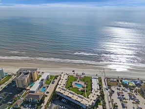 Vue aérienne d’un hôtel situé sur la côte, composé de plusieurs bâtiments, avec une piscine et une plage.