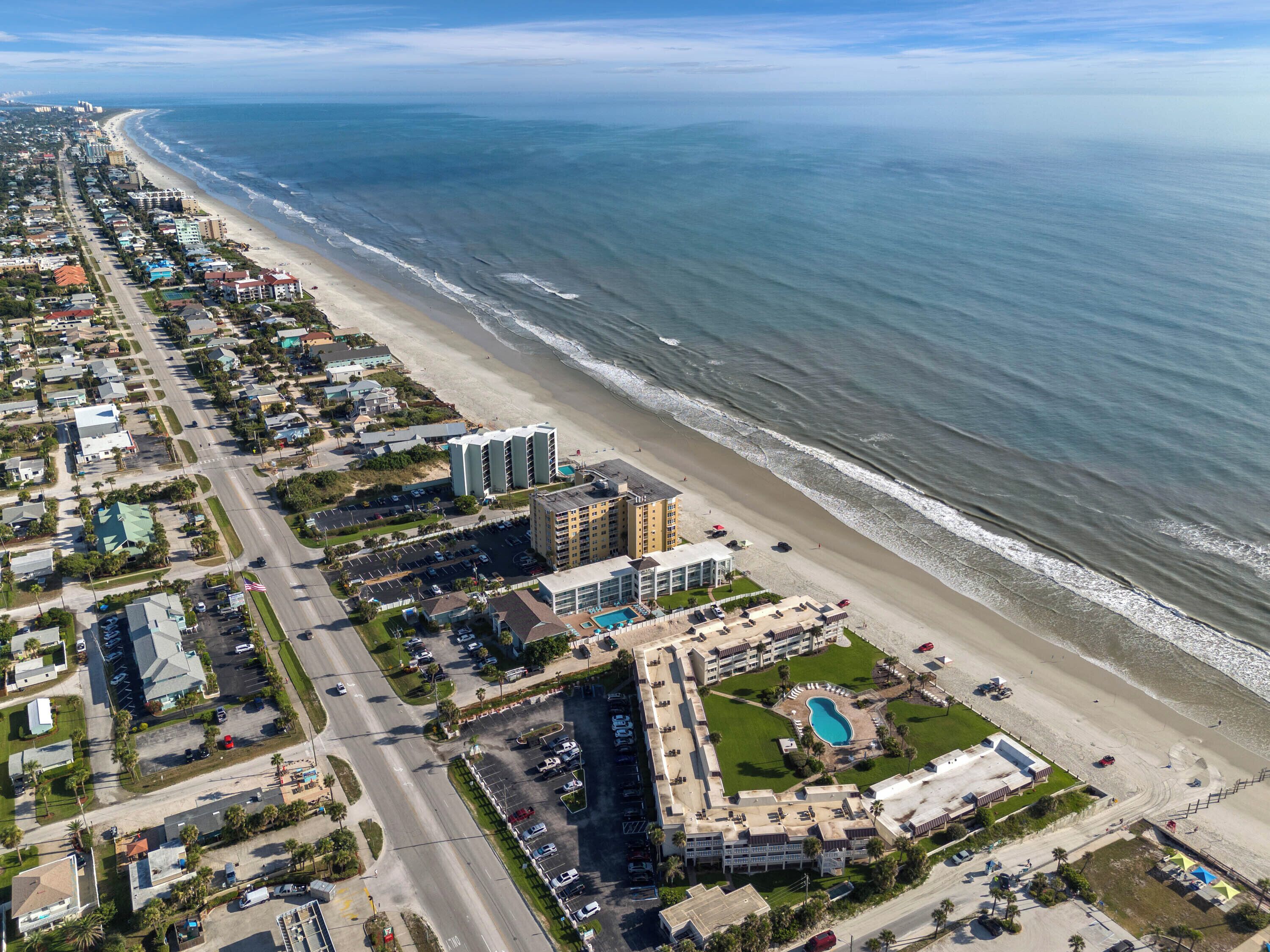 An aerial view of a coastal hotel with a beachfront, parking lots, and a swimming pool.