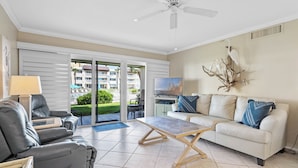 A modern living room with a white sofa, grey recliner, and a wooden coffee table. There is a ceiling fan, a TV, and a decorative bird sculpture on the wall. - Chateau by the Sea 13A Pure Ocean Bliss! (New Smyrna Beach)