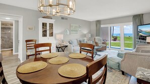 A spacious living room with a large window offering a view of the outdoors, a dining area with a wooden table and chairs, and a modern chandelier.