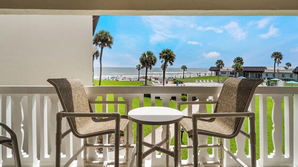 A balcony with two chairs and a small table, overlooking a beach with palm trees and a clear sky. - Chateau by the Sea 25C Serenity by the Sea (New Smyrna Beach)