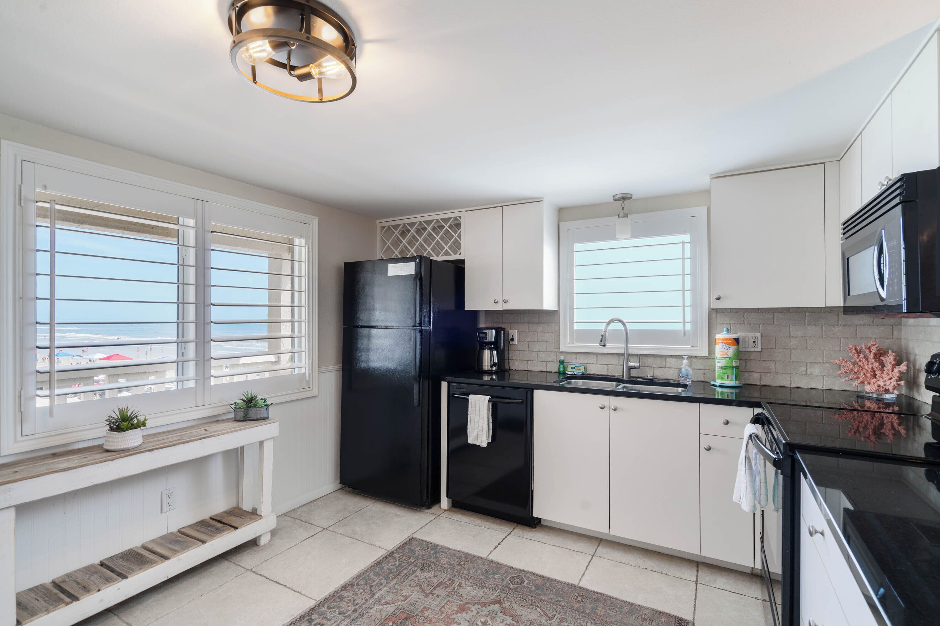 Cocina moderna con armarios blancos, un refrigerador negro, microondas y cafetera. Hay dos ventanas con persianas, un banco con plantas y una alfombra en el suelo.