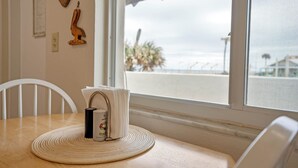 Ein Holztisch mit einer gewebten Tischmatte, ein weißer Stuhl und ein Fenster mit Blick auf den Strand und Palmen.
