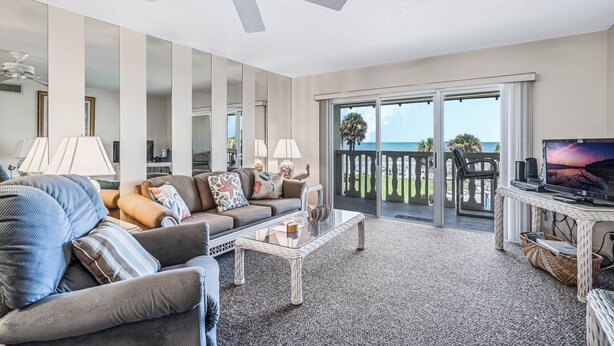 Ein großzügiges Wohnzimmer mit einer großen Couch, einem Couchtisch und einem Fernseher auf einem Schrank. Eine Glastür verschiebt sich zu einem Balkon mit Blick aufs Meer.