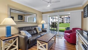 A living room with a sofa, coffee table, and a red chair. There is a ceiling fan, a large mirror, and a sign that reads "BEACH HOUSE". - Chateau by the Sea 12B Ocean View Retreat! (New Smyrna Beach)