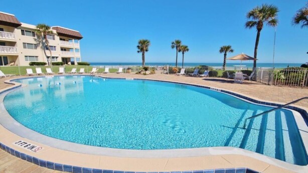 A swimming pool with a tiled edge, surrounded by lounge chairs and palm trees, with a building in the background. - Chateau by the Sea 12B Ocean View Retreat! (New Smyrna Beach)