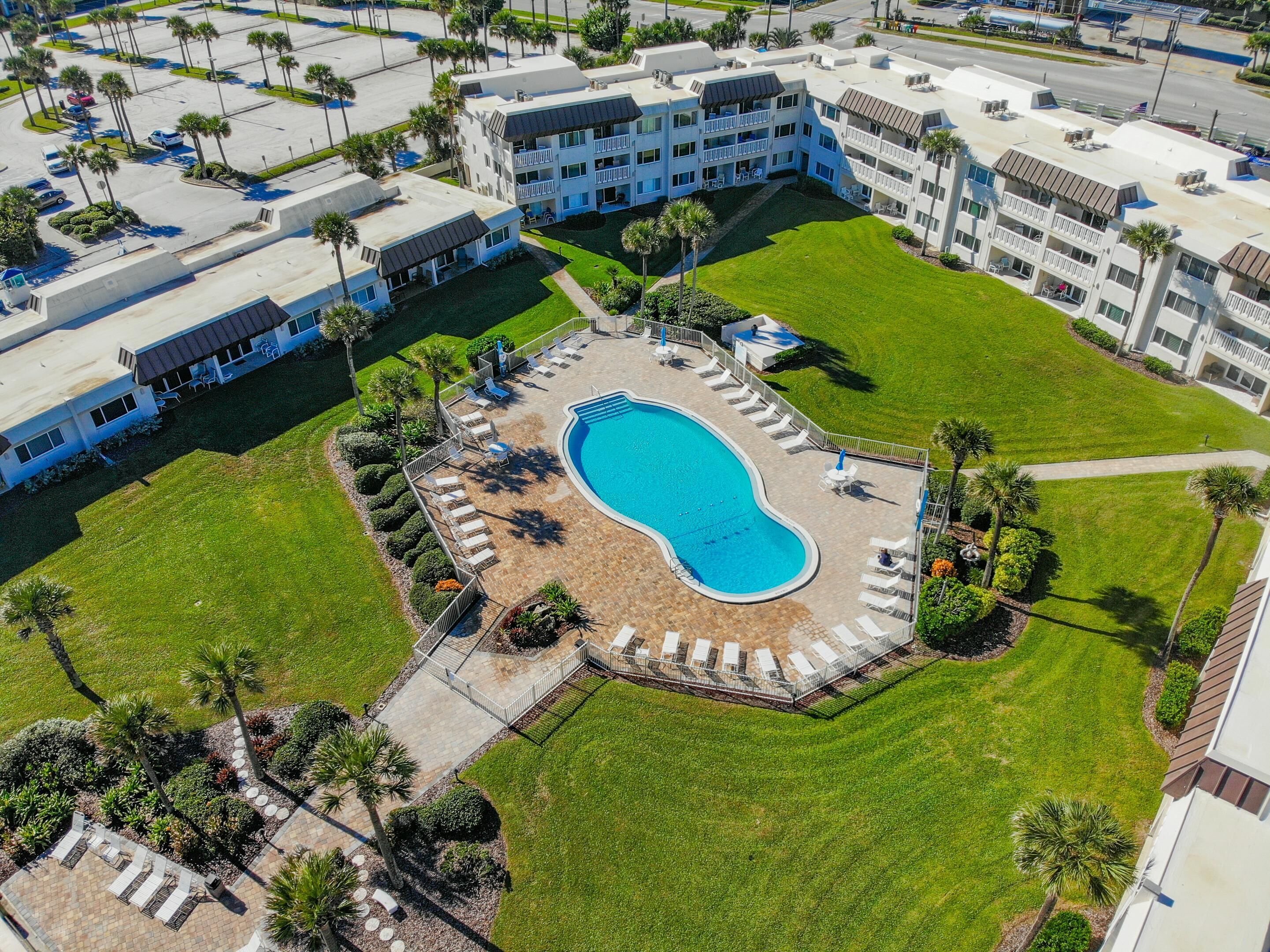 Vista aérea de un hotel con piscina rodeada por una terraza, palmeras y un césped cuidado.
