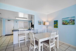 Un comedor con una mesa y sillas blancas, una pequeña cocina con refrigerador, y una puerta que da a otra habitación.