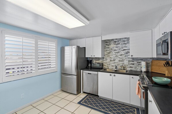 Una cocina moderna con armarios blancos, electrodomésticos de acero inoxidable, un respuesto de azulejos y una ventana con persianas.