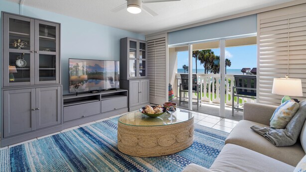 Ein Wohnzimmer mit Fernseher, einem Glastisch und einer Couch. Ein Balkon bietet Blick auf den Ozean.